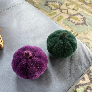 Two fuzzy pumpkins, one purple and one green, on a textured surface.